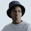 Young man wearing a dark bucket hat and light gray shirt against a light background.