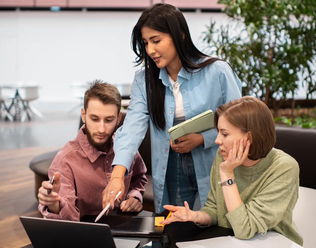Three young professionals collaborating over a laptop in a modern office setting, with one standing and pointing at the screen.