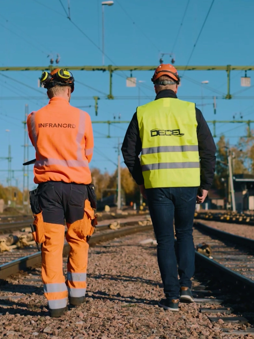 En installatör och en Decel-medarbetare i varselkläder går längs spåren på en rangerbangård under kontaktledningar.