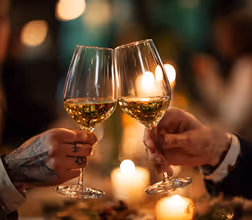 Two hands clinking glasses of white wine in a warm, candlelit setting.