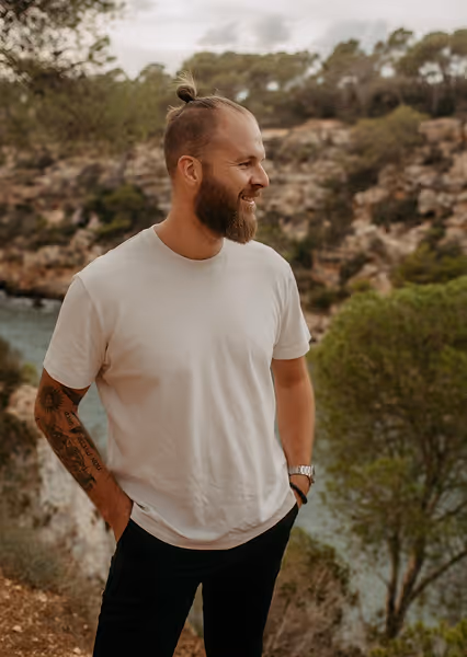 Bearded man with a top knot hairstyle, wearing a white t-shirt and black pants, standing outdoors near a river and trees.