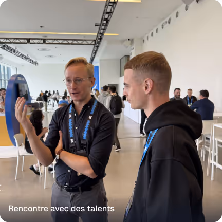 Two men in conversation inside a modern, open conference space with people in the background.