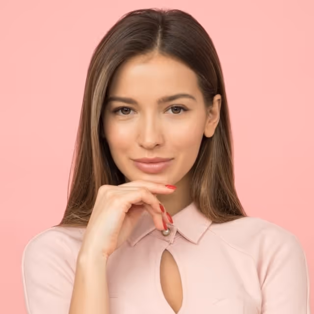 Young woman with long brown hair, wearing a pale pink top, resting her chin on her hand against a pink background.