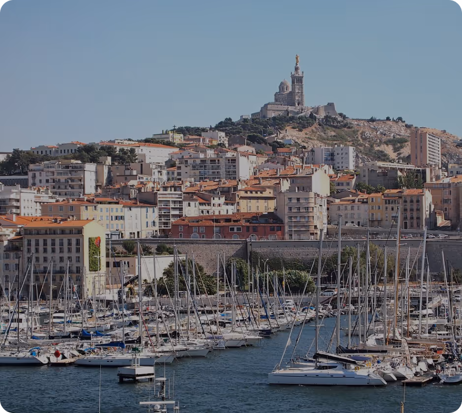 Port de plaisance avec de nombreux voiliers amarrés devant des bâtiments urbains et une colline surmontée d'une grande basilique à Marseille.