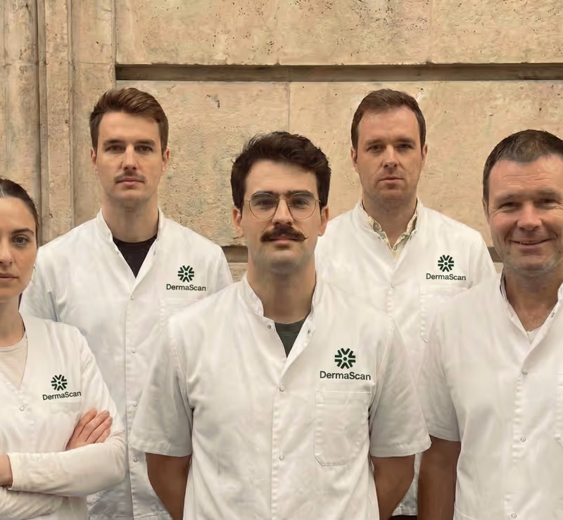 Groupe de professionnels en blouses blanches avec logo DermaScan, debout devant un mur en pierre.