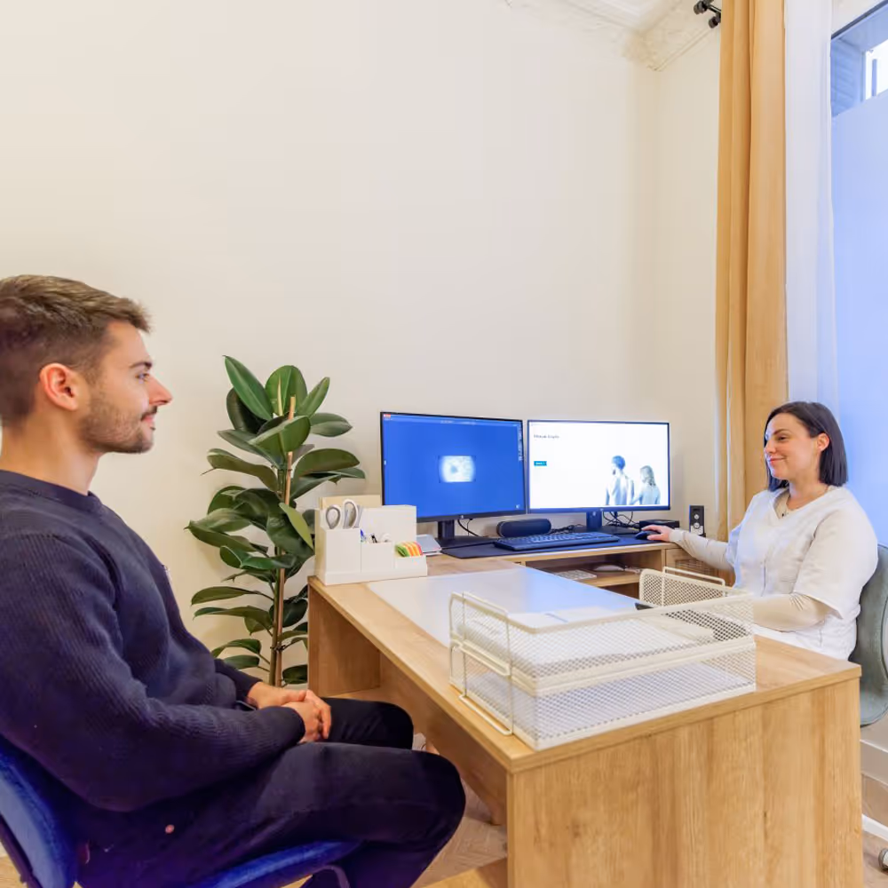Un homme et une femme discutent dans un bureau moderne avec deux écrans d'ordinateur sur un bureau en bois.