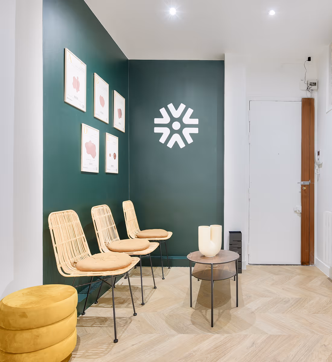 Salle d'attente moderne avec trois chaises en rotin sur coussins beiges, murs verts foncés décorés de cadres et un logo blanc, table basse ronde et un pouf jaune.