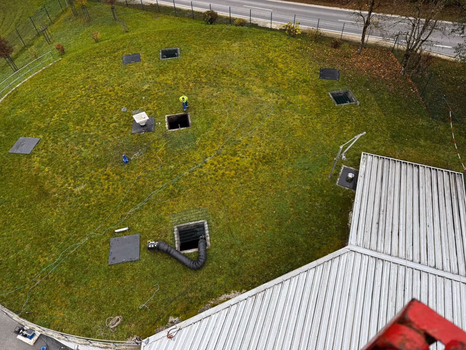 Luftaufnahme eines großen Grasfeldes mit mehreren offenen Bodenschächten, einem Arbeiter in Schutzkleidung und verschiedenen Schläuchen und Kabeln, neben einem Gebäude mit Metall-Dach.