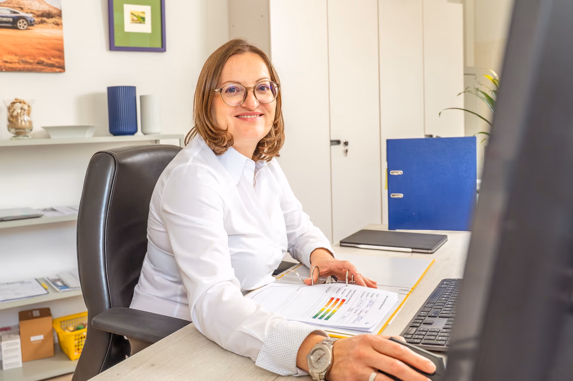 Frau mit Brille und weißem Hemd sitzt am Schreibtisch und lächelt, während sie am Computer arbeitet.
