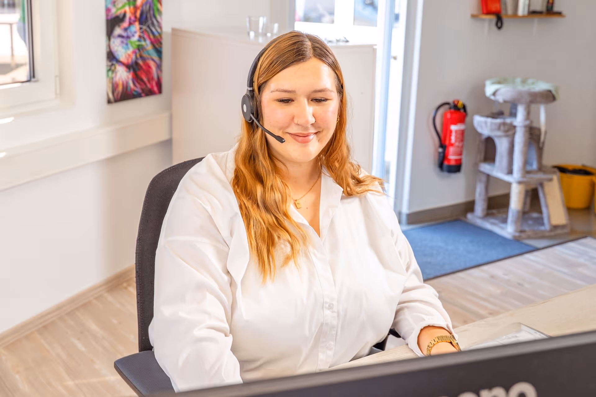 Lächelnde Frau mit Headset sitzt an einem Schreibtisch und arbeitet an einem Computer in einem hellen Büro.