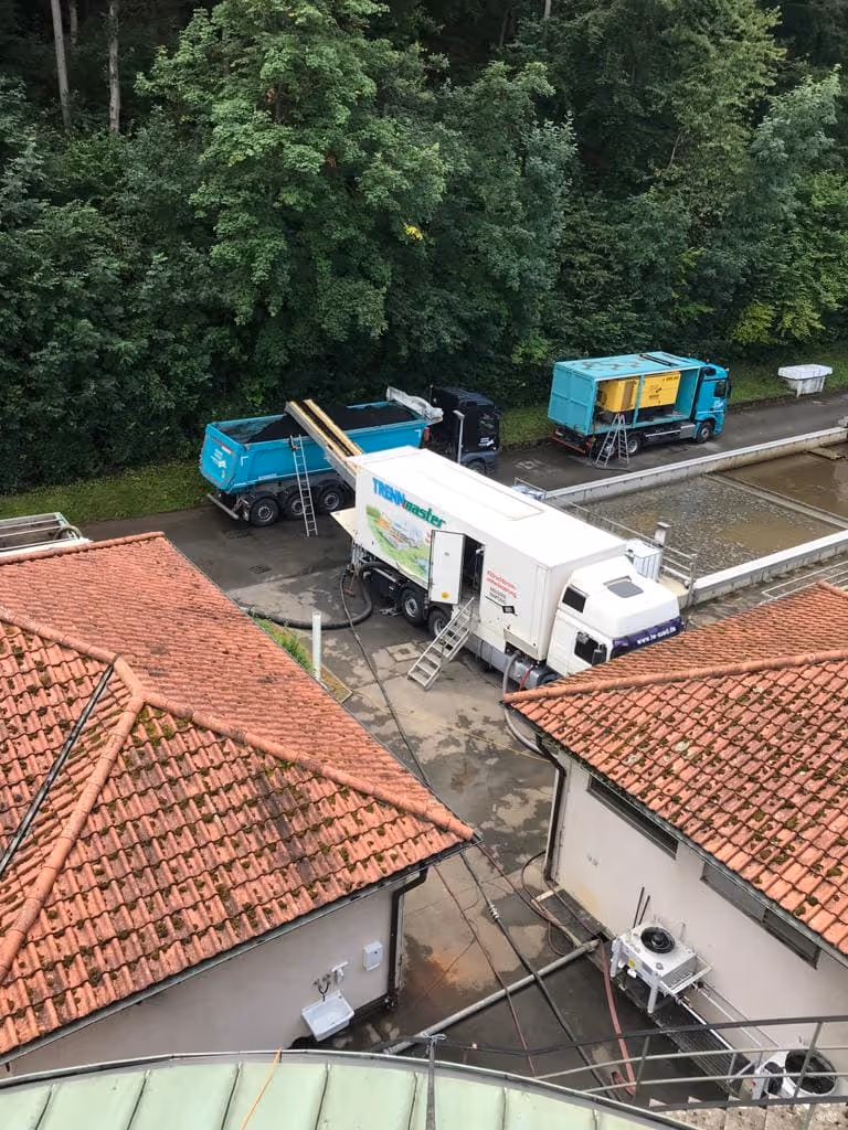 Bau- oder Reinigungsfahrzeuge, darunter ein weißer Truck mit der Aufschrift ‚TRENMaster‘, stehen auf einem gepflasterten Hof neben Gebäuden mit roten Dächern und einem bewaldeten Hintergrund.