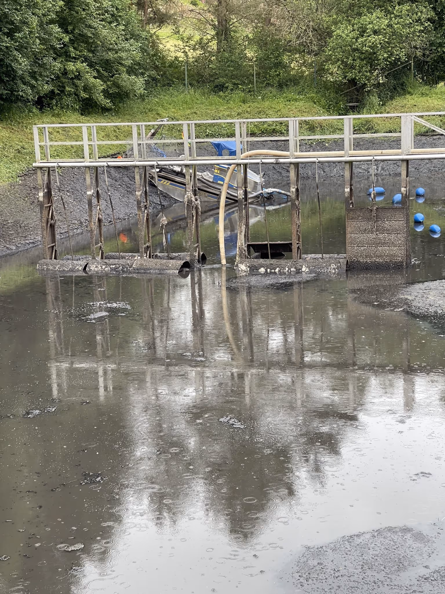 Wasserbehandlungsanlage mit einem Metallgeländer, verschmutztem Wasserbecken und blauen Schwimmern, umgeben von Gras und Bäumen.