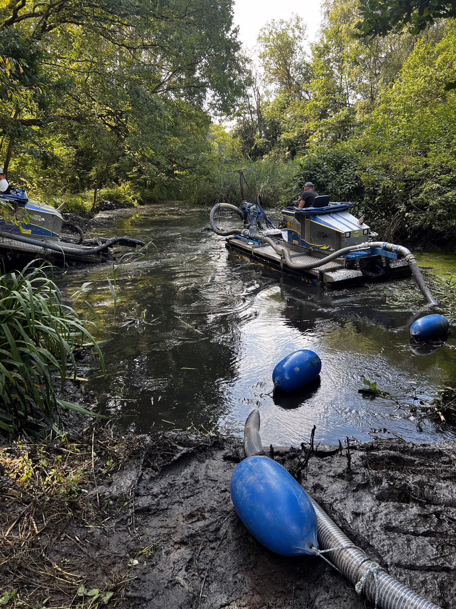 Maschine auf einem Fluss mit Schläuchen und blauen Schwimmkörpern zur Reinigung oder Entschlammung, umgeben von dichtem Grün.