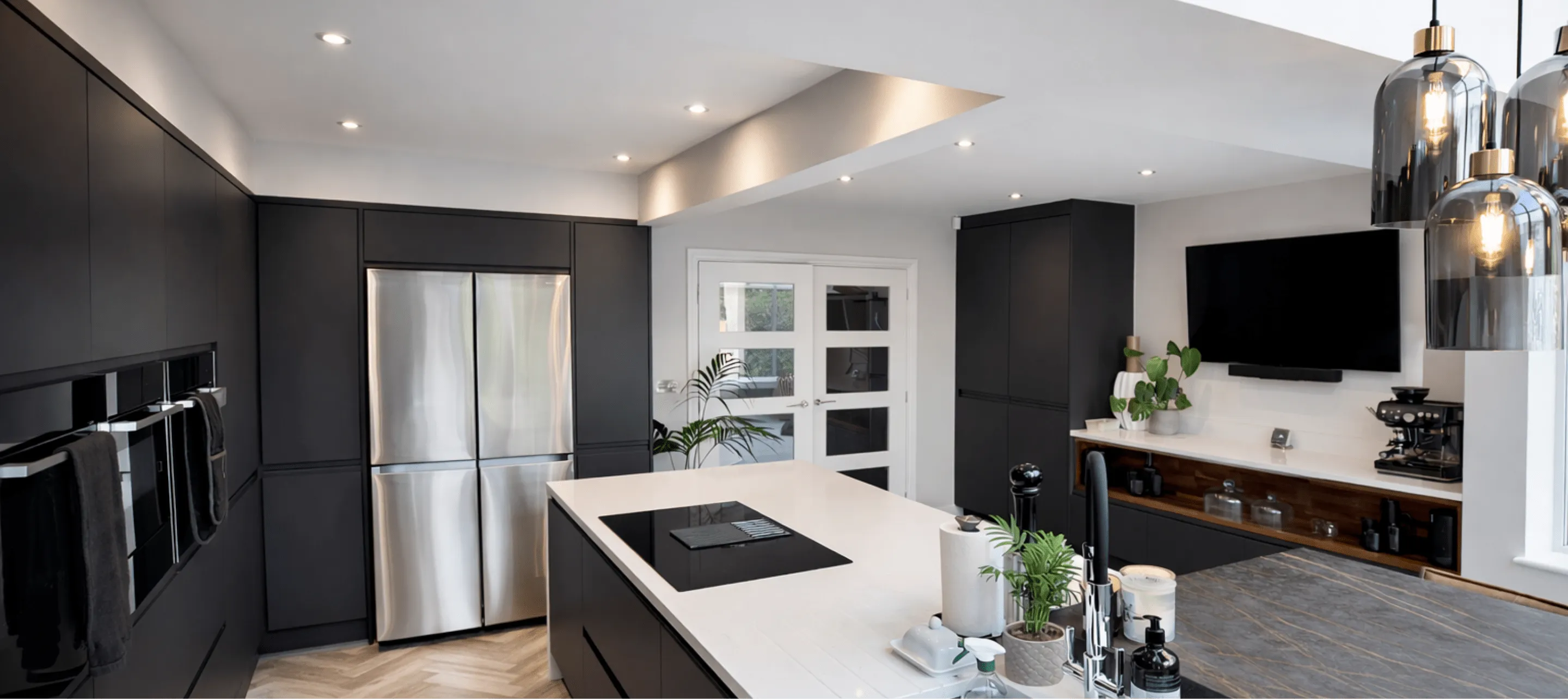 Modern kitchen with black cabinetry, stainless steel fridge, white island countertop, hanging pendant lights, and a wall-mounted TV.