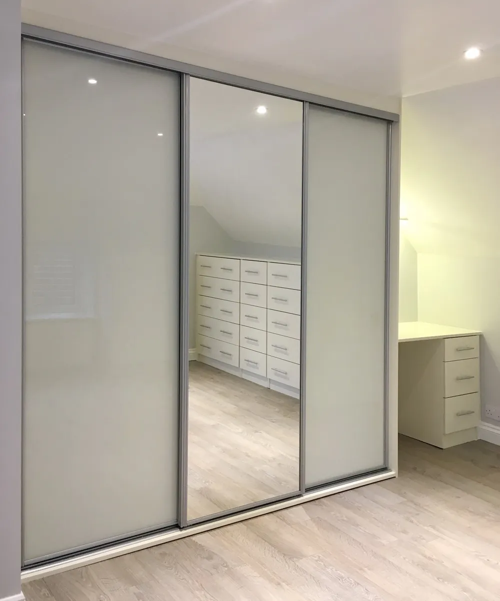 White sliding wardrobe with one mirrored door reflecting a set of white drawers and light wood flooring.