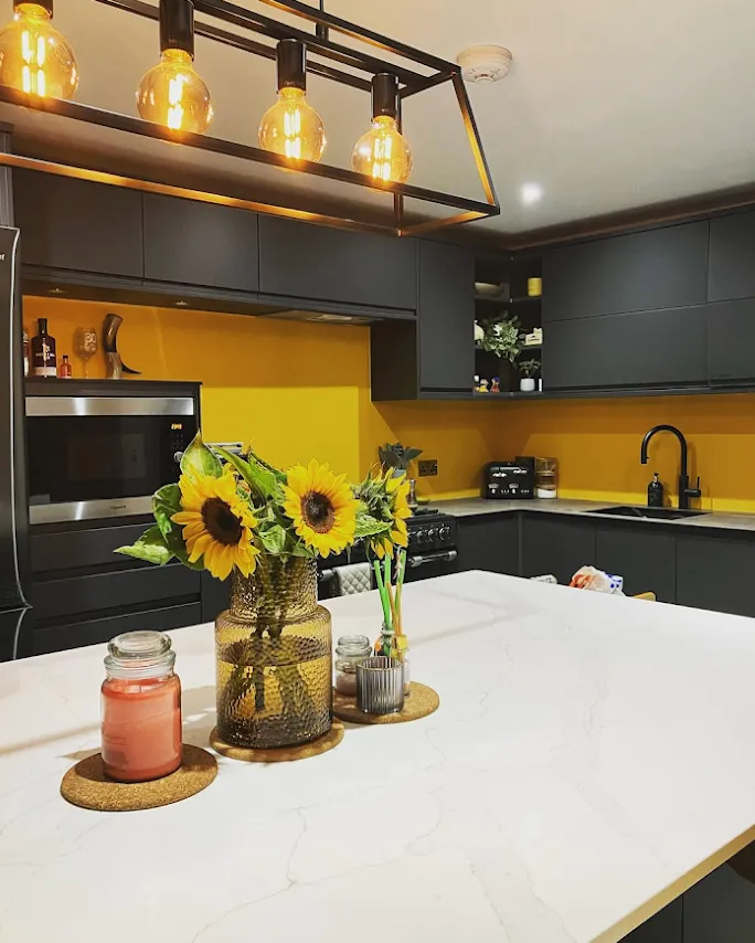 Modern kitchen with dark cabinets, yellow backsplash, white marble countertop island, sunflowers in glass vase, and hanging Edison bulb lights.