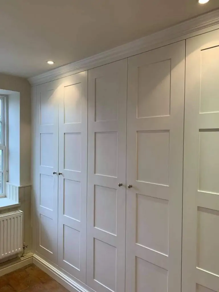 Built-in white paneled wardrobes with silver knobs illuminated by recessed ceiling lights next to a window and radiator in Rotherham
