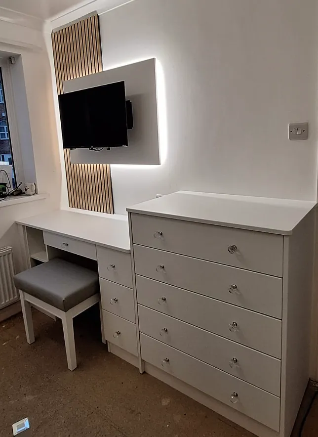 Modern white bedroom furniture set with a chest of drawers and a vanity desk with a stool, a wall-mounted TV, and vertical wooden slat decor with backlighting.