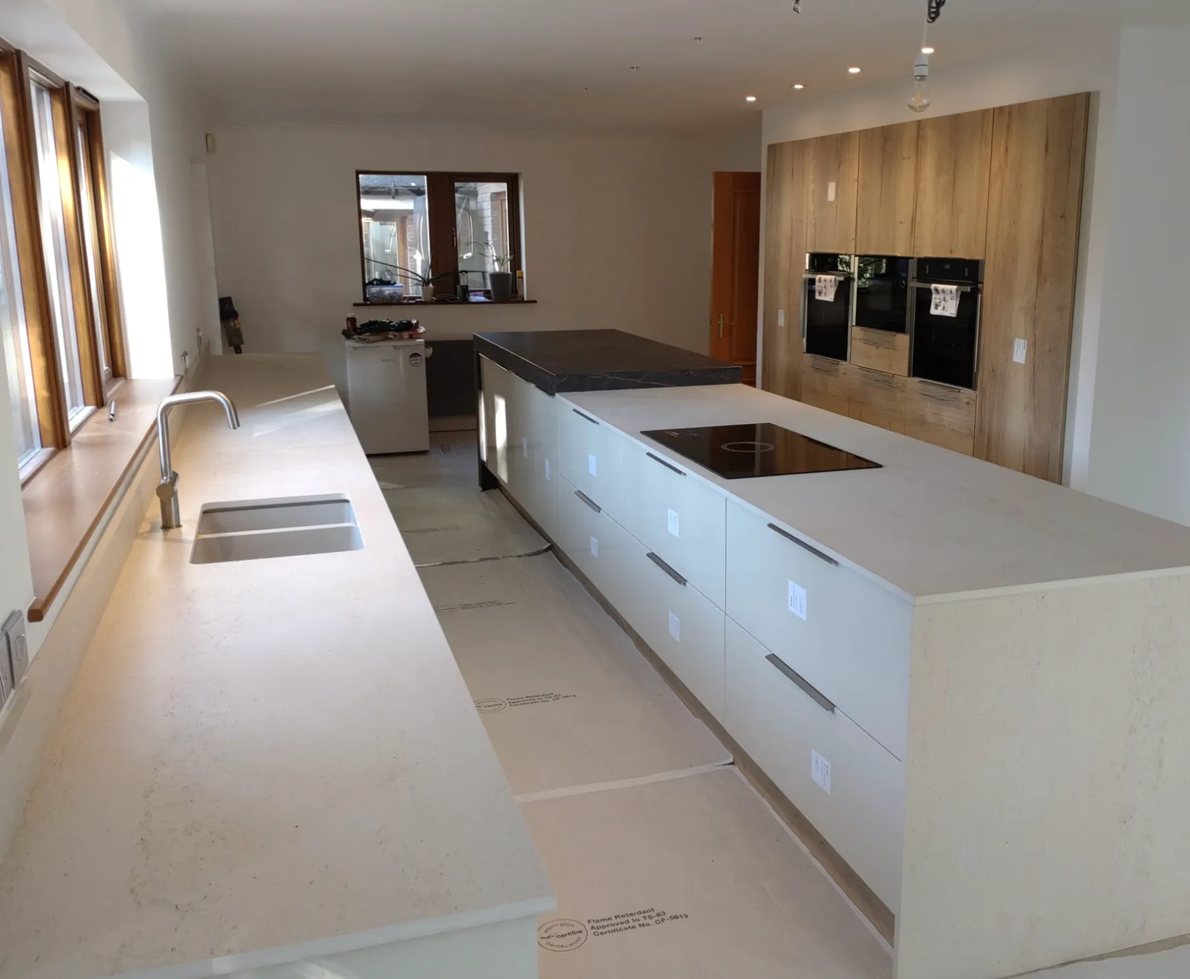 Modern kitchen under construction with white countertops, built-in stovetop on island, stainless steel sink, wooden framed windows, and built-in ovens in wood cabinetry.