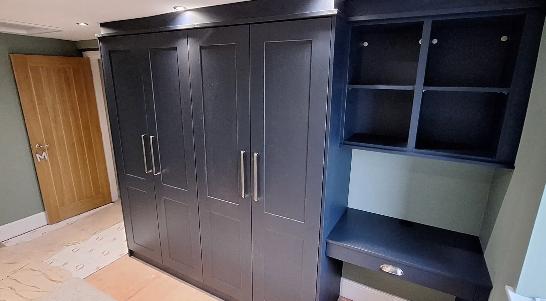 Dark blue built-in wardrobe with four doors and adjacent desk with shelves in a room with green walls and a wooden door.