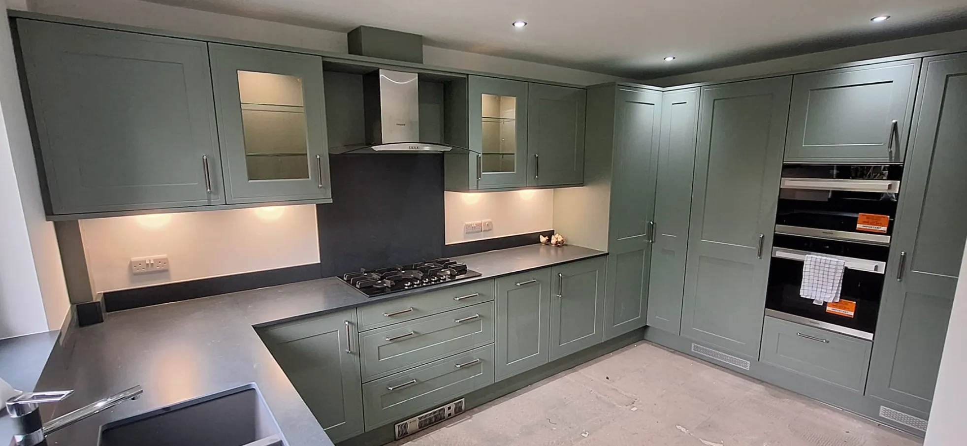 Modern kitchen with matte green cabinetry, built-in gas cooktop, stainless steel hood, double oven, and dark gray countertops.