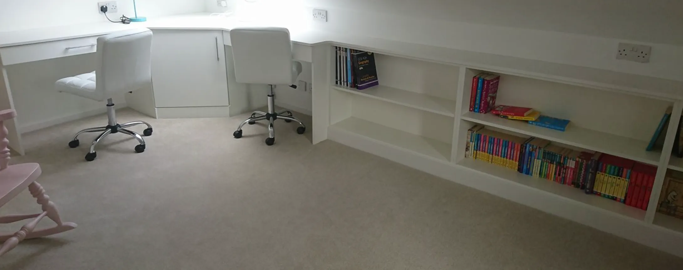 Corner study area with two white swivel chairs, white built-in desk, and shelves with colorful books.