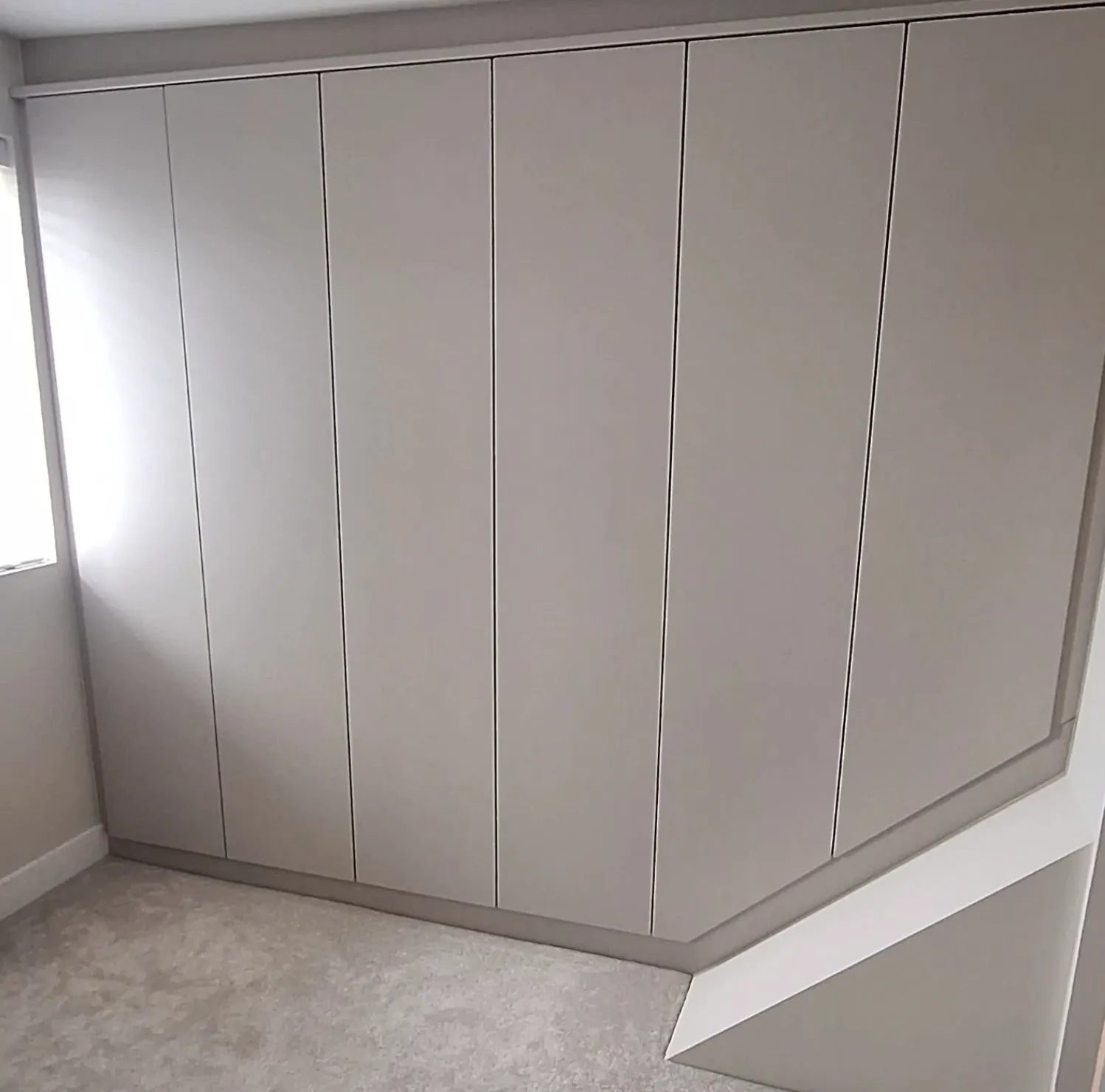 Built-in angled wardrobe with six tall, smooth cashmere doors in a carpeted room with a window on the left in Penistone