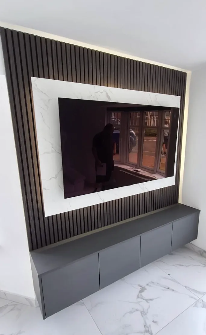 Modern wall-mounted flat-screen TV framed by white marble paneling and vertical dark wood slats above a sleek gray floating cabinet on a white marble floor.
