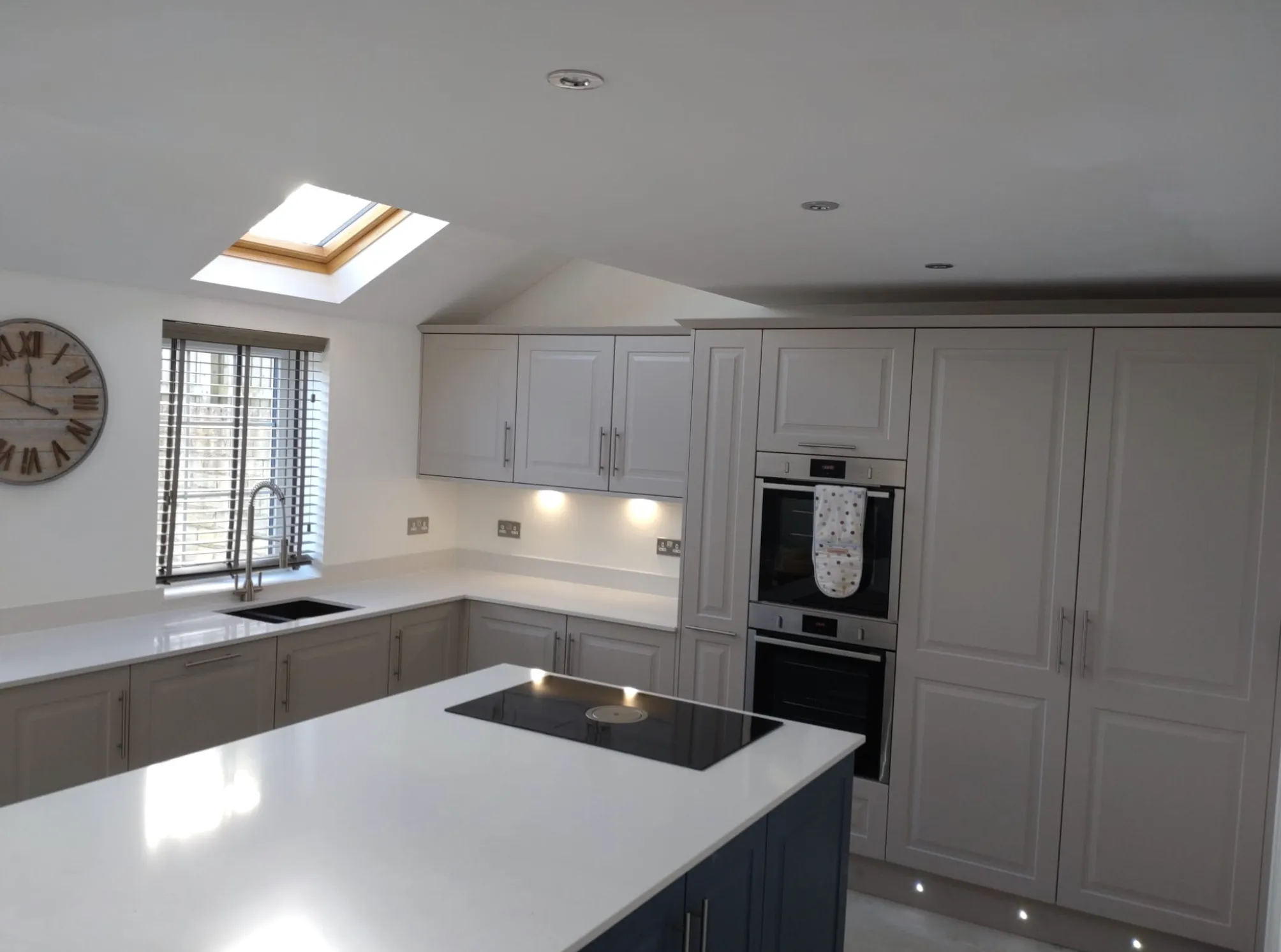 Modern kitchen with light gray cabinets, white countertops, island with black cooktop, skylight, and wall clock in Rotherham