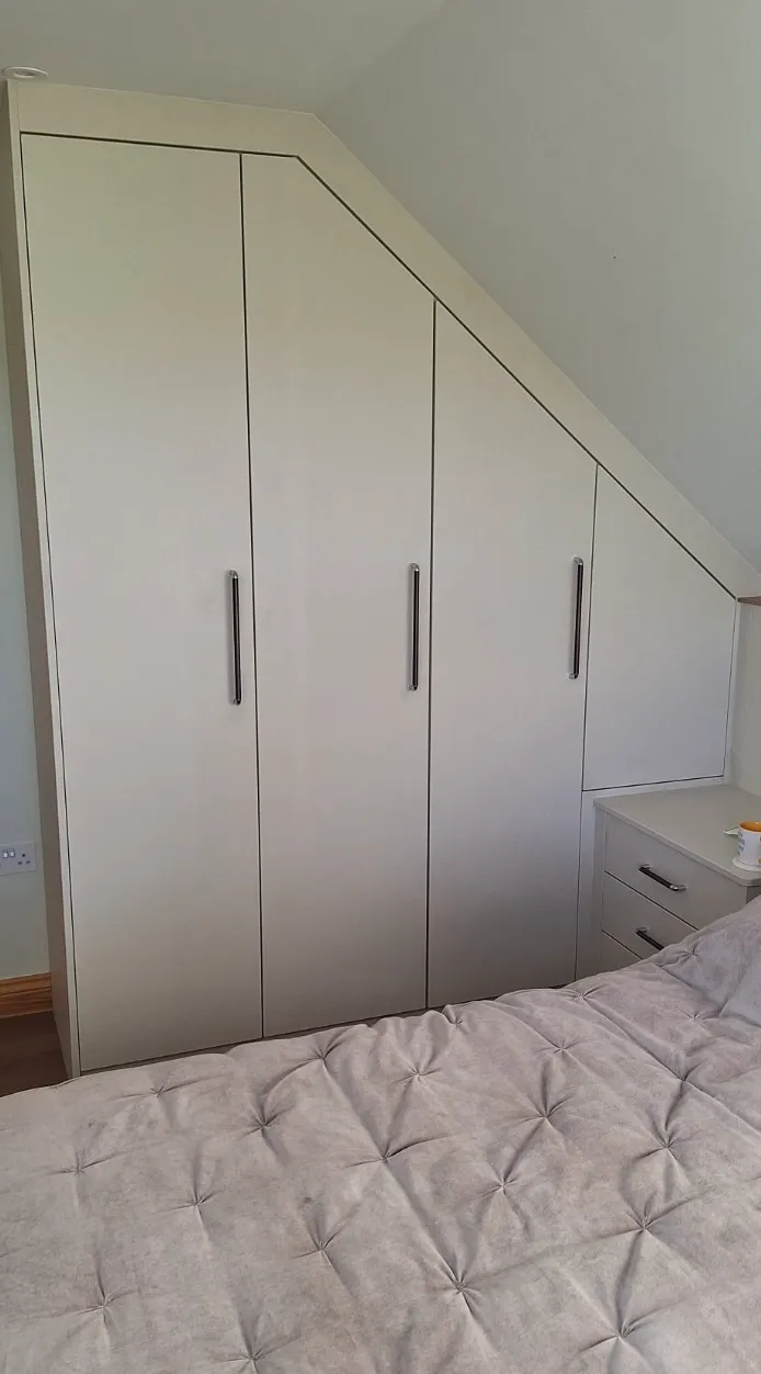 Built-in wardrobe with three tall doors and one smaller door under a sloped ceiling, next to a bedside table with drawers and a bed with a tufted gray quilt.