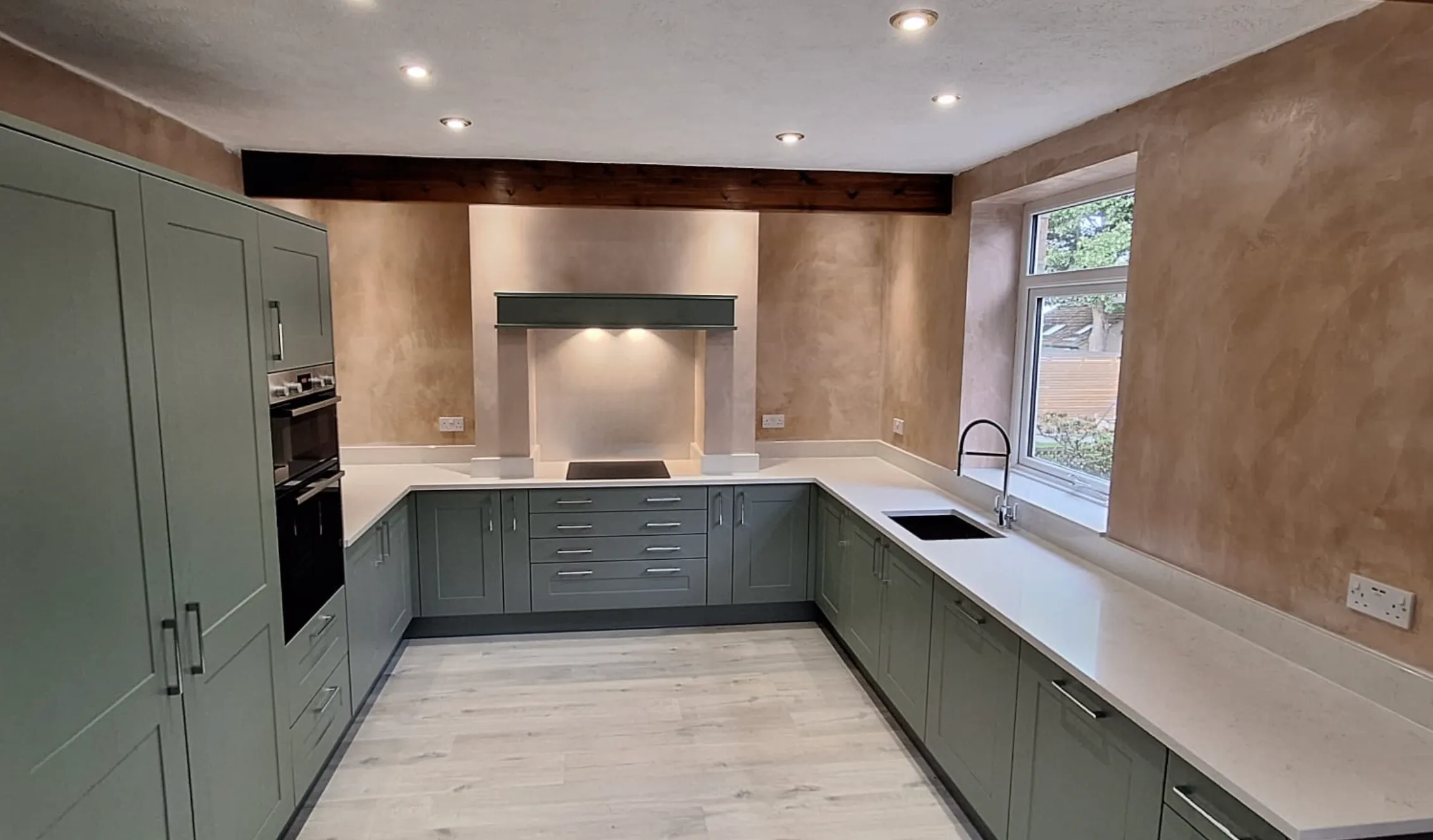 Modern U-shaped kitchen with matte green cabinets, white countertops, built-in ovens, black sink under window, and recessed ceiling lights in Barnsley 