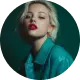 Woman with short blonde hair wearing a teal shirt and red lipstick against a dark background.