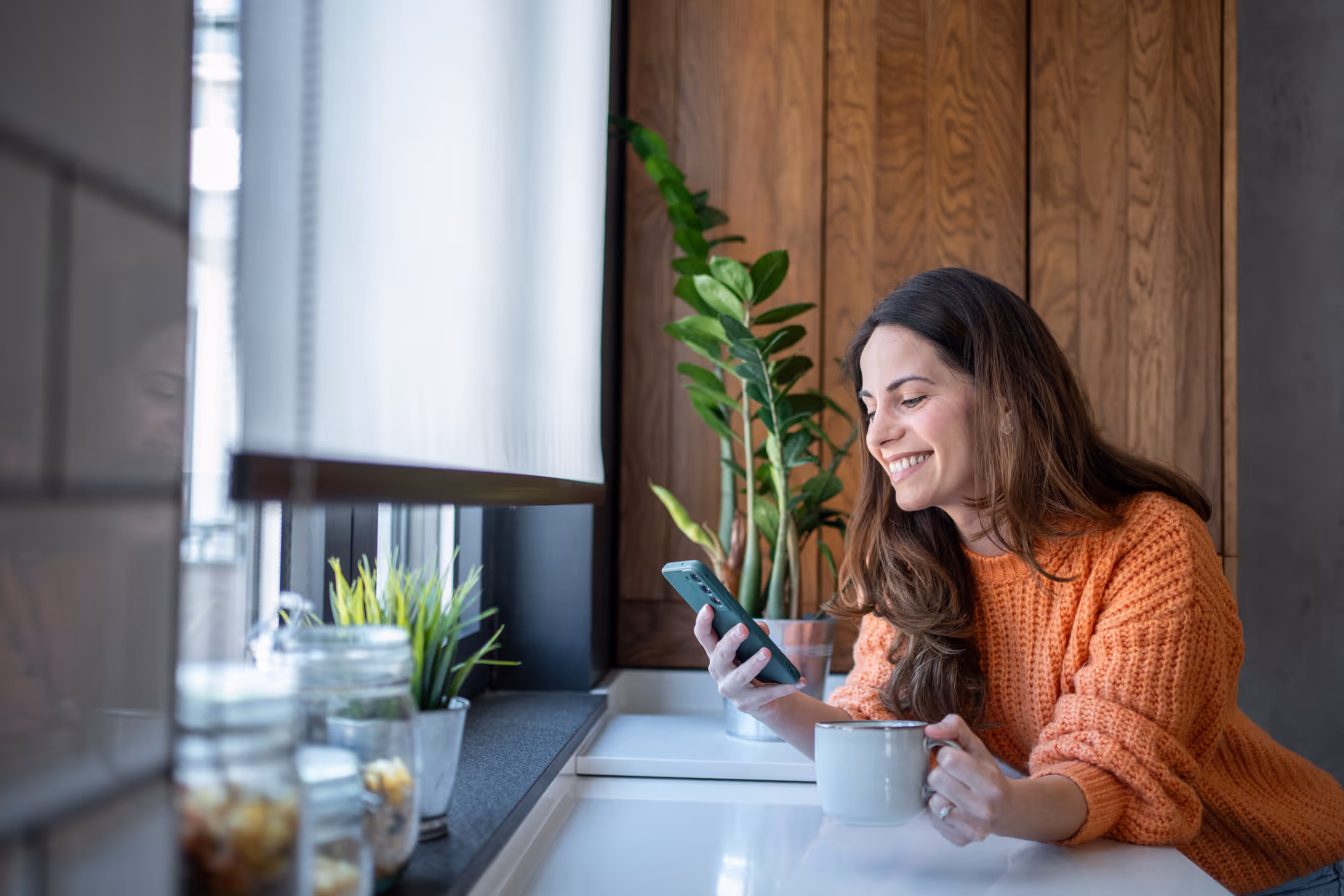 Smilende kvinde i orange sweater ser på sin telefon og holder en kop ved et køkkenbord.