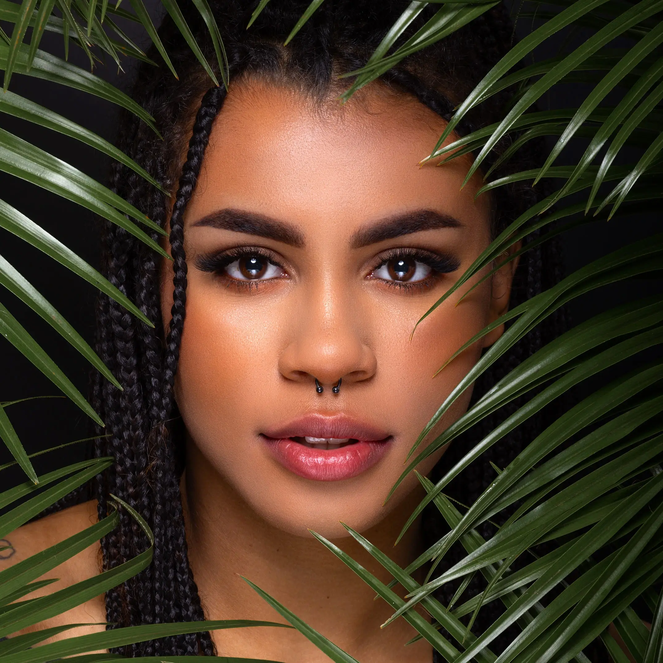 Portret młodej kobiety z warkoczami i kolczykiem w nosie, otoczonej zielonymi liśćmi roślin.