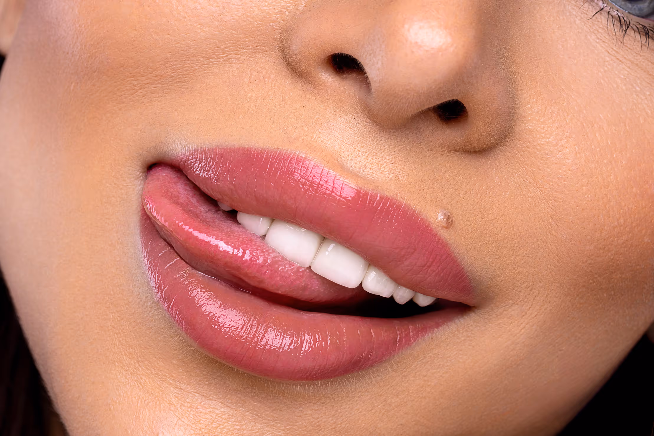 Zbliżenie na usta z różowym połyskującym makijażem, język dotyka górnej wargi, widoczny drobny pieprzyk na skórze.