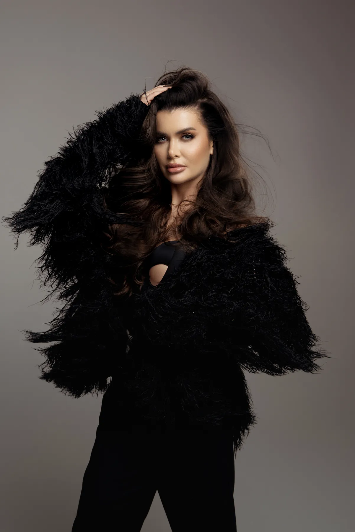 Kobieta z długimi, ciemnobrązowymi włosami, ubrana w czarny puszysty sweter, pozująca na szarym tle.
