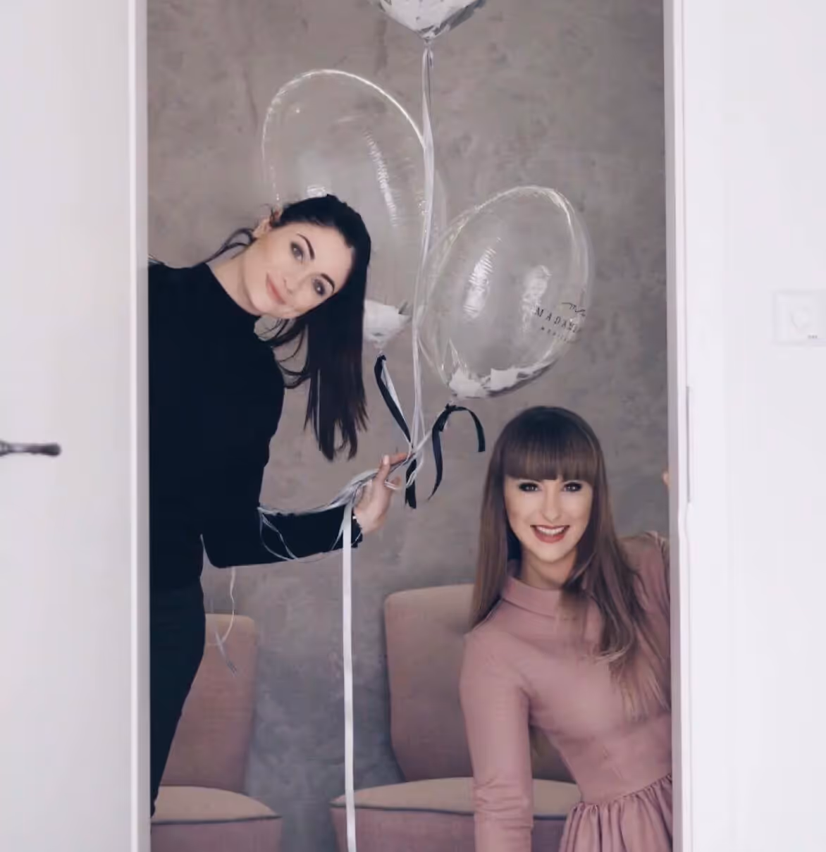 Dwie uśmiechnięte kobiety w drzwiach, jedna trzyma przezroczyste balony, druga odstaje ścianę.