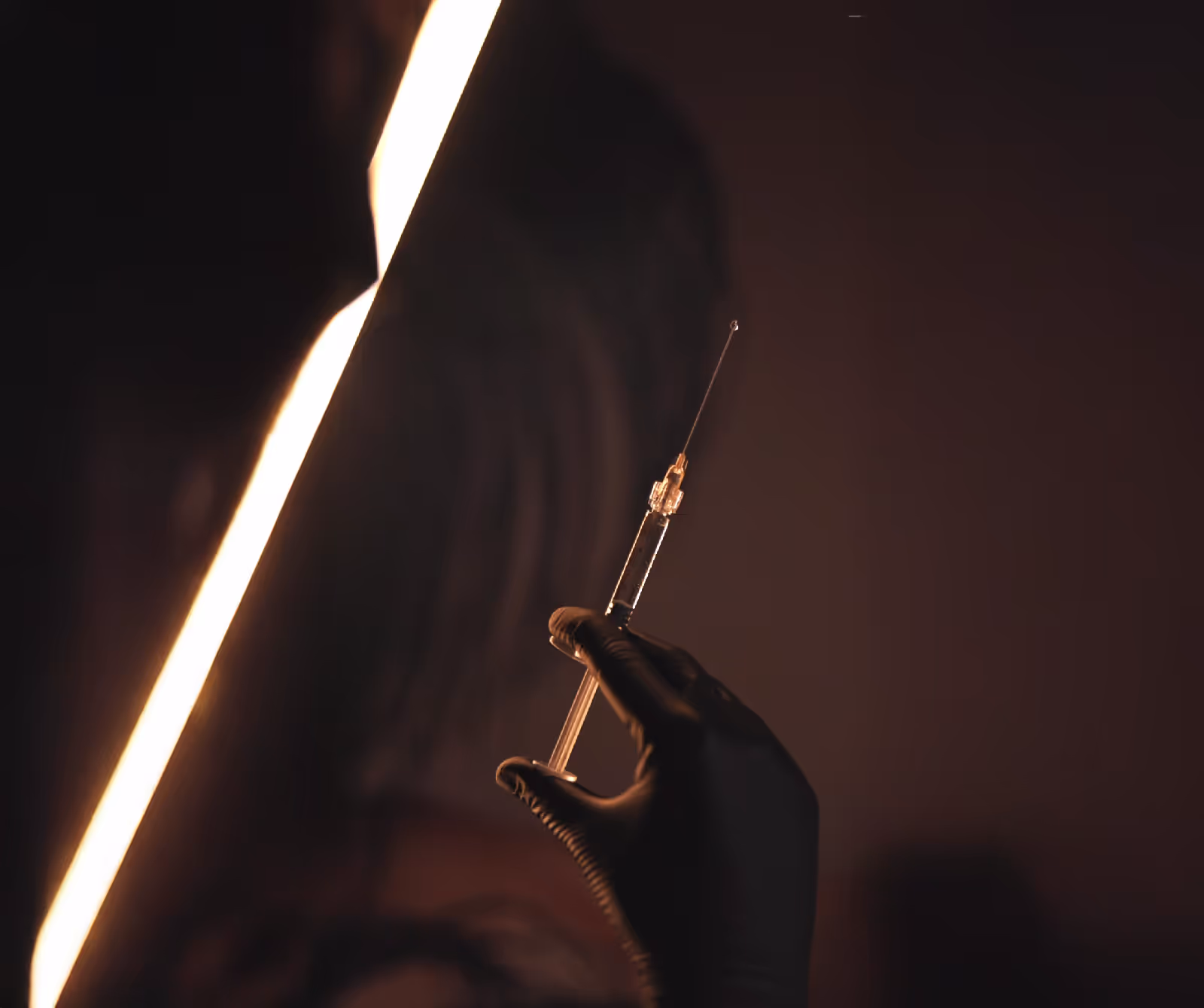 Ręka w czarnej rękawiczce trzymająca strzykawkę z kroplą na końcu igły na ciemnym tle.