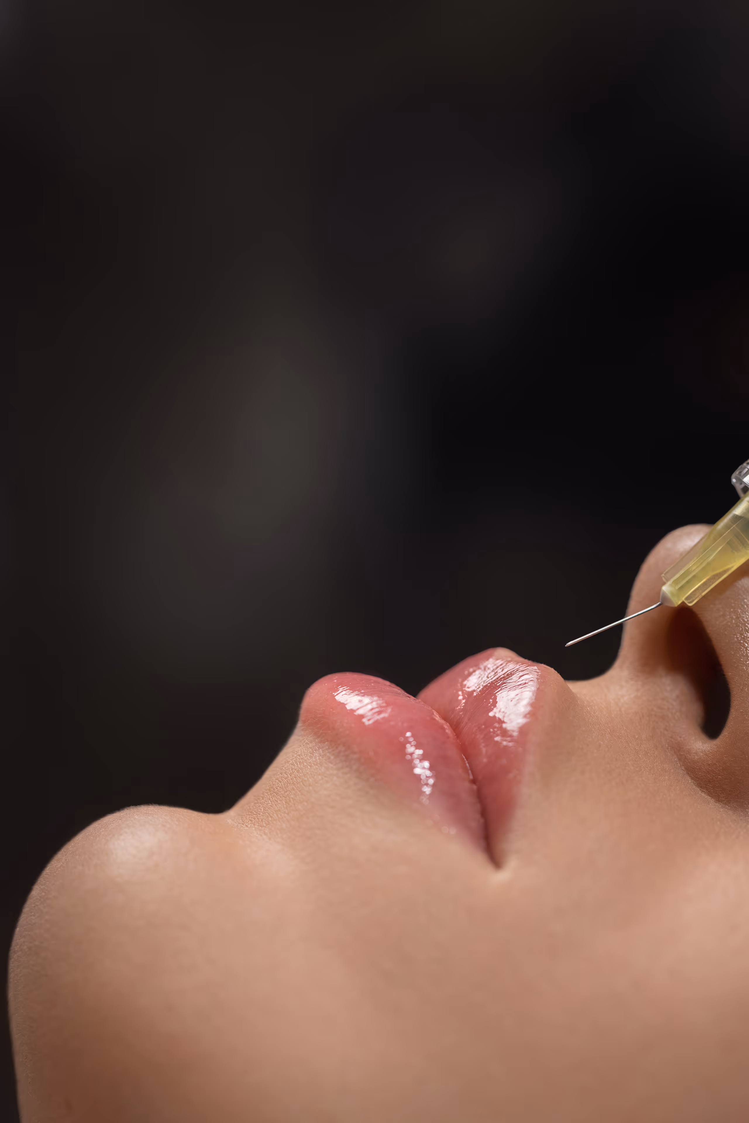 Bliska pionowa fotografia ust z połyskującą pomadką i igły strzykawki przygotowanej do iniekcji.