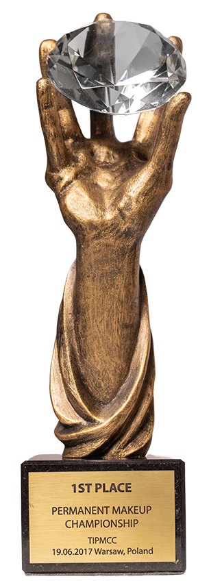 Złota statuetka przedstawiająca dłoń trzymającą duży przezroczysty kryształ, z tabliczką na podstawie z napisem o pierwszym miejscu w Permanent Makeup Championship w Warszawie 2017.