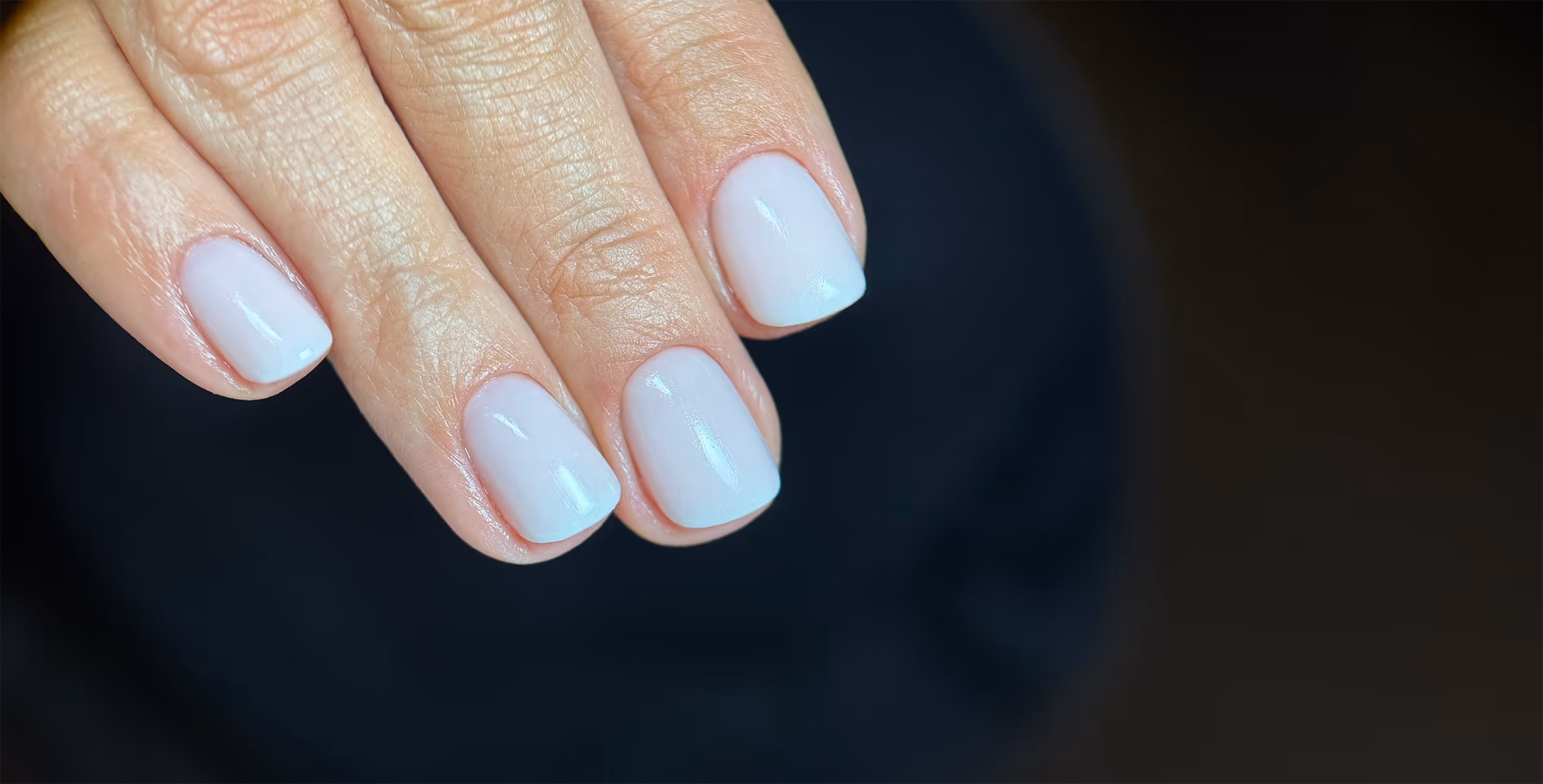 Kobiece dłonie z krótkimi, białymi paznokciami o gładkim manicure na ciemnym tle.