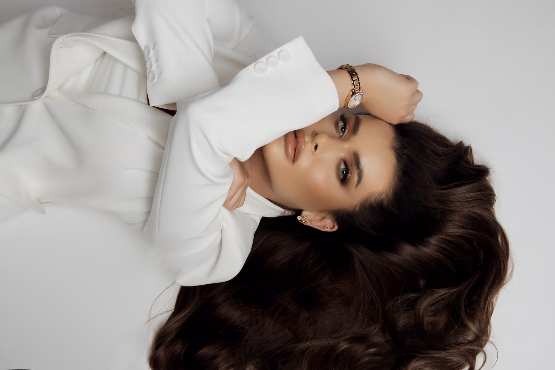 Kobieta w białej marynarce leży na ziemi, podpierając się ręką na czole, z rozpuszczonym, falującym ciemnym włosami.