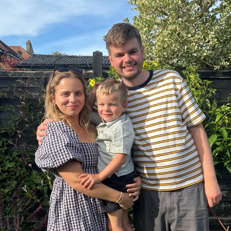 Smiling woman in a checkered dress and man in a striped t-shirt holding a young boy in a garden on a sunny day.