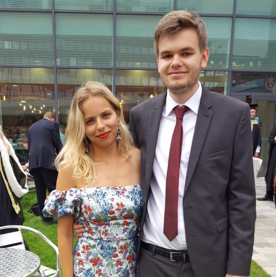 Young couple standing on grass outside a modern glass building, woman wearing floral off-shoulder dress, man in dark suit with red tie.