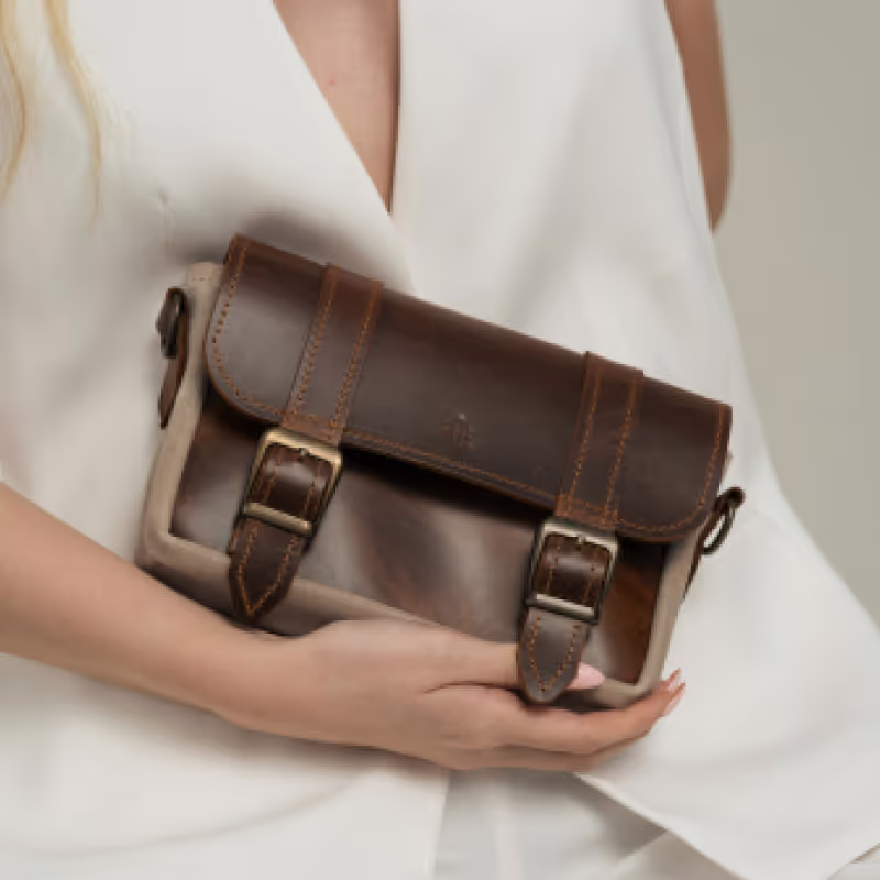 Woman wearing white holding a small brown leather satchel with two buckle straps.