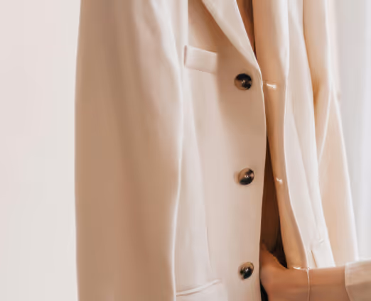 Close-up of a person wearing a cream-colored blazer with black buttons and a beige sweater underneath.