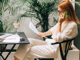 Woman in white outfit sitting on a chair, holding a document and talking on a green phone, with a table and a plant in the background.
