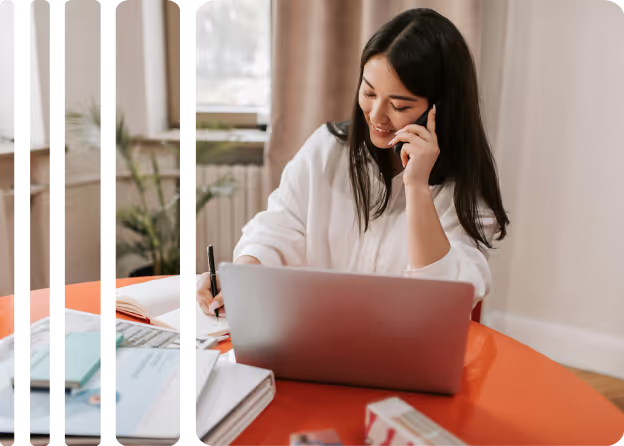 Woman sitting at a desk with an open laptop, writing in a notebook, and talking on a smartphone.
