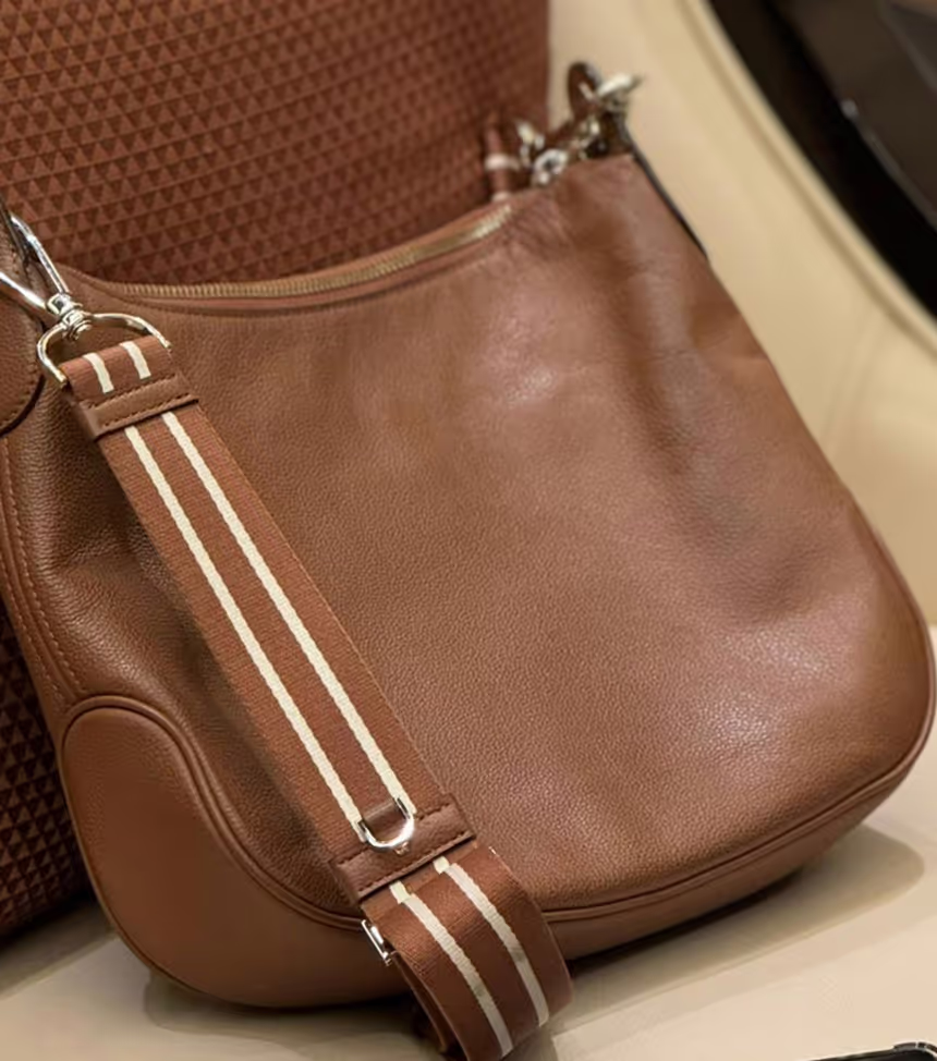 Brown leather shoulder bag with a striped fabric strap resting on a beige and brown textured surface.