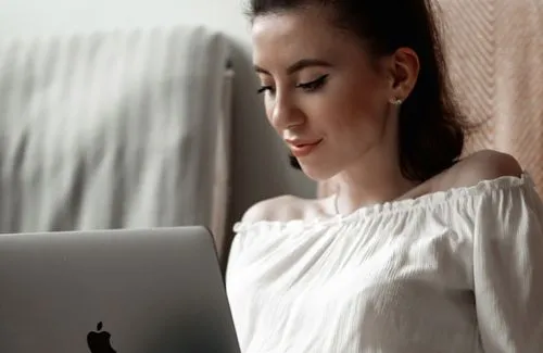 Young woman in a white off-shoulder top looking at a laptop screen with a slight smile.